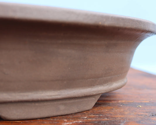 Close-up of a brown ceramic bonsai pot showing its textured clay body and flared rim, resting on a wooden surface against a light blue background.