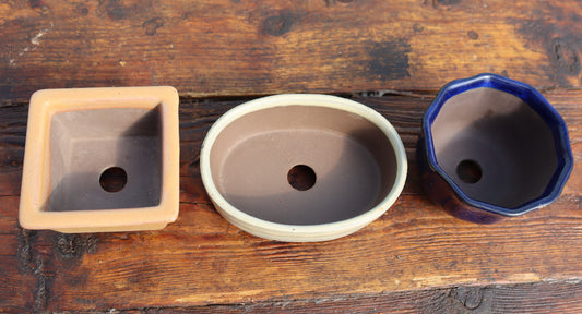 Three ceramic bonsai pots with drainage holes displayed side-by-side on a rustic wooden surface. The pots include a square terracotta pot, an oval beige pot, and an octagonal dark blue glazed pot.