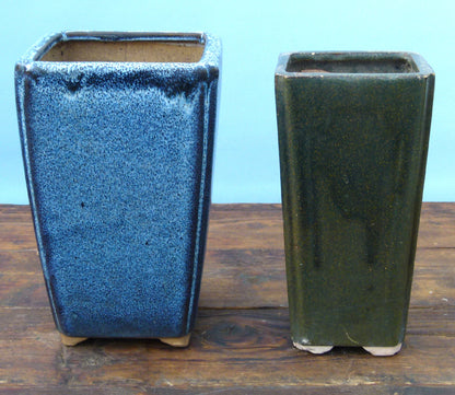 Two square ceramic pots, one blue and one green, on a wooden surface with a light blue background.