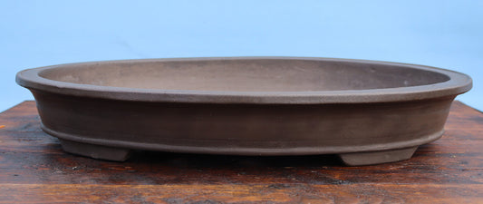 An oval ceramic bonsai pot in a dark brown matte finish, displayed on a wooden surface against a light blue background.