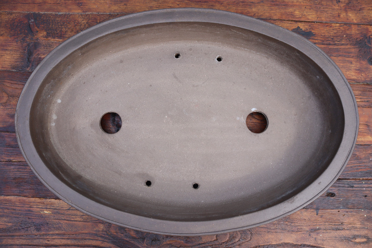 An oval-shaped dark gray ceramic bonsai planter base with drainage holes, shown from a top-down view on a rustic wooden background.