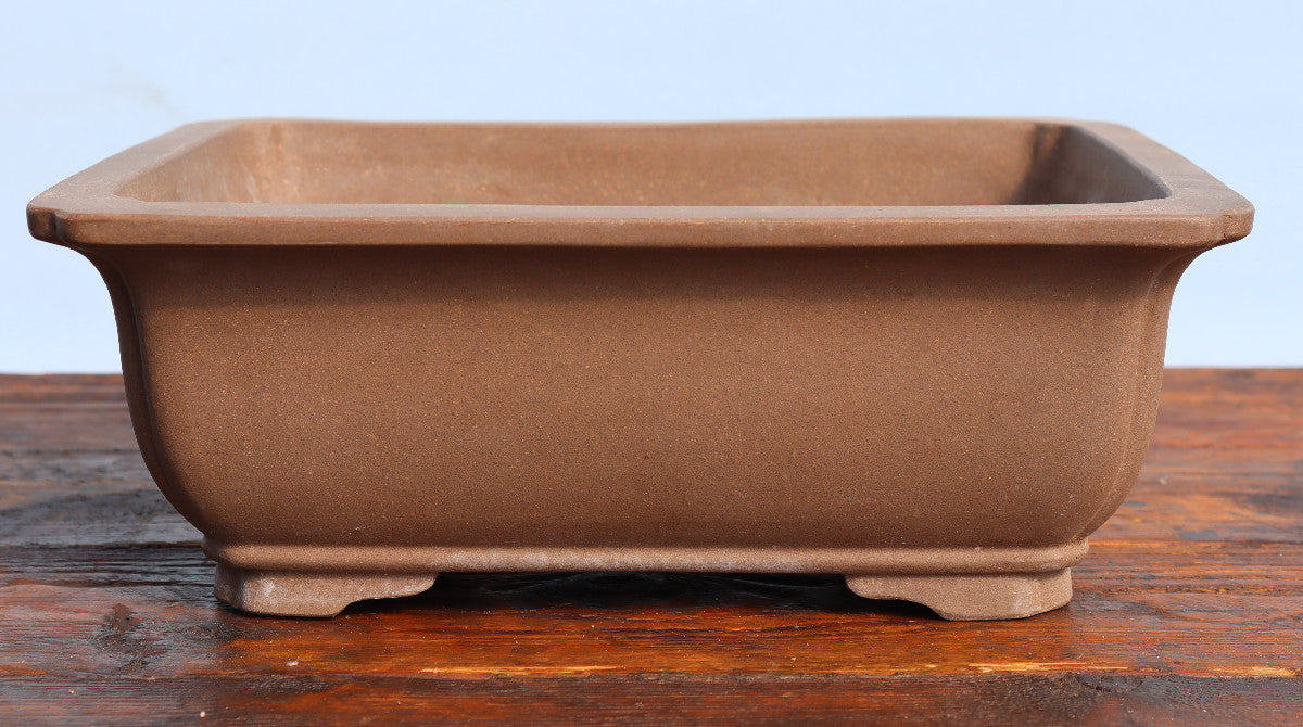 A rectangular brown ceramic bonsai pot with a matte finish, shown on a wooden surface against a light blue background.