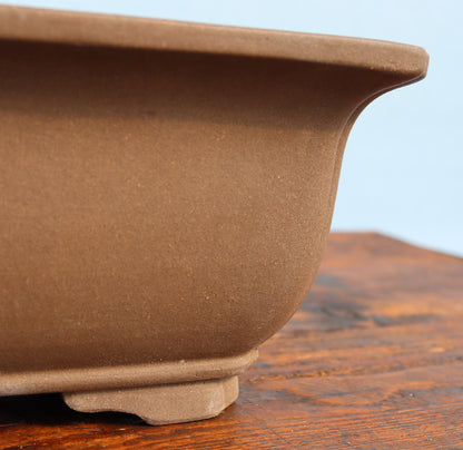Close-up of a brown ceramic bonsai pot showing its textured matte glaze and footed base, resting on a wooden surface against a light blue background.