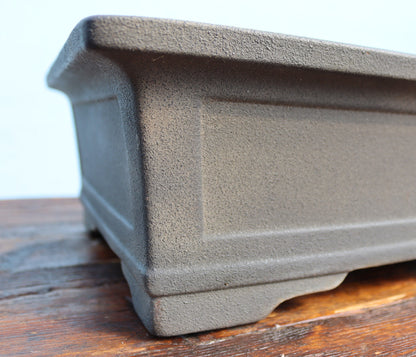 Close-up of a rectangular dark gray cast-iron or concrete bonsai planter resting on a rustic wooden surface against a light background.