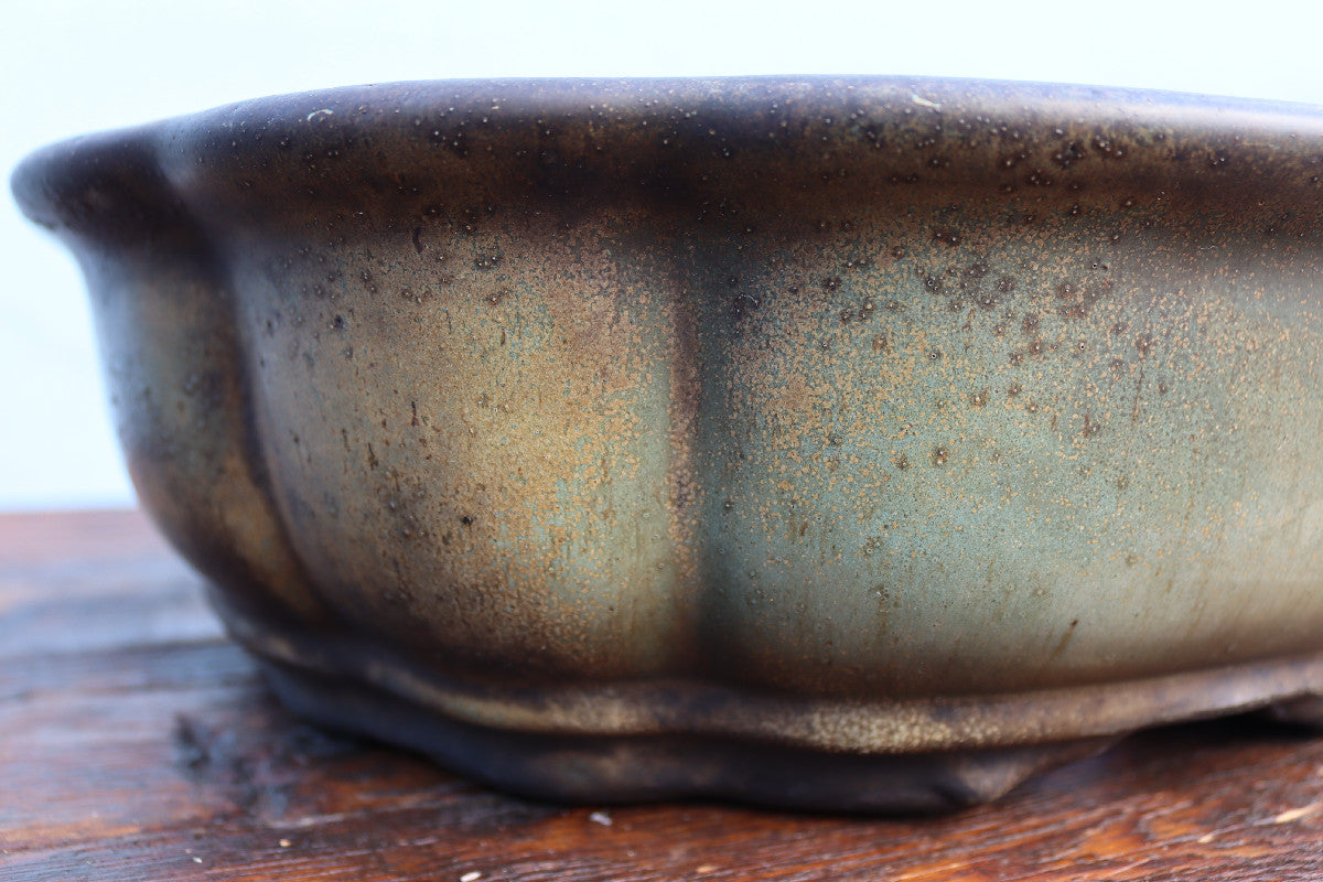 Close-up of a ceramic planter pot with a textured, aged glaze showing brown and muted green tones, sitting on a wooden surface.